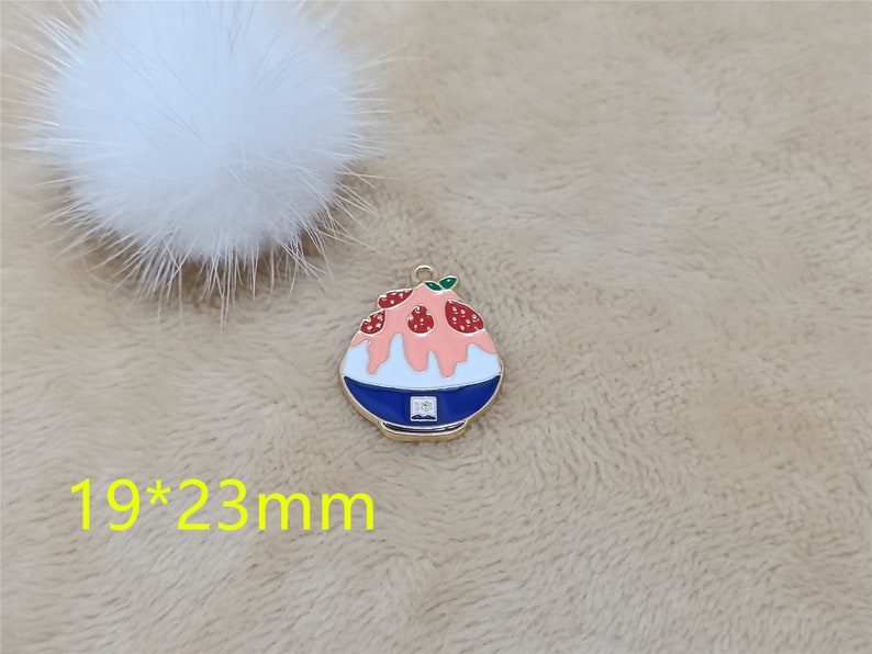 May include: A gold-toned metal charm shaped like a bowl of shaved ice with pink and white frosting and red strawberries. The charm is 19 mm by 23 mm.