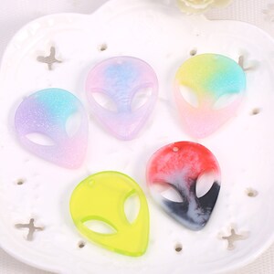 以下が含まれることがあります： 紫、青、黄、赤、黒の色合いの、カラフルなエイリアンヘッドペンダントが5つ。各ペンダントの上部には、紐を通すための穴があります。ペンダントは、グラデーション効果のある半透明の素材で作られています。