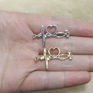 Puede incluir: Dos encantos de plata y oro en forma de corazón con un estetoscopio dentro. Los encantos están en una mano.