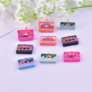 May include: Assortment of eight colorful cassette tape charms with various designs, including palm trees, mountains, and waves. Each charm has a silver loop for attaching to jewelry or other crafts.