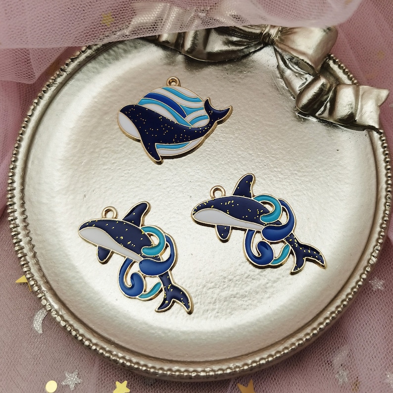 May include: Three gold-tone metal whale charms with blue and white enamel paint. The whales have a sparkly finish and are perfect for jewelry making.