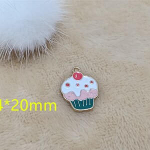 May include: A gold-colored charm shaped like a cupcake with white frosting, pink icing, and red sprinkles. The cupcake is 14mm by 20mm.