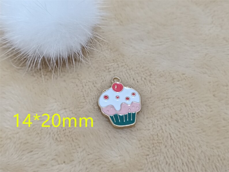 May include: A gold-colored charm shaped like a cupcake with white frosting, pink icing, and red sprinkles. The cupcake is 14mm by 20mm.