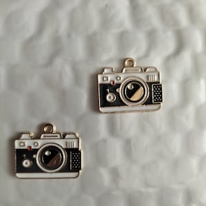 May include: Two gold-colored metal camera charms with black and white enamel details. The cameras have a round lens with a black center and a white rim. The charms have a loop at the top for attaching to a necklace or bracelet.