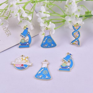 Op de afbeelding: Zes blauwe en witte emaille hangers met wetenschappelijke thema's. De hangers omvatten een microscoop, een bekerglas, een DNA-streng, een bekerglas met het woord "Chemie" erop en twee andere bekerglazen.