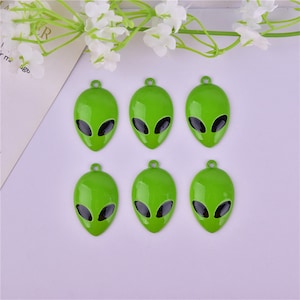 May include: Six green alien face charms with black eyes. The charms are made of metal and have a shiny finish.