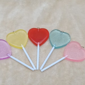 Può includere: Cinque lecca-lecca a forma di cuore su bastoncini. I lecca-lecca sono di colore viola, giallo, rosso, blu e rosa.