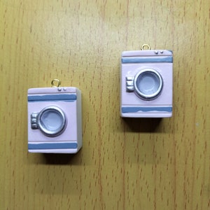 May include: Two miniature pink and gray washing machine ornaments with a silver-colored door and a gold loop for hanging.