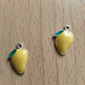 May include: Two silver charms shaped like yellow mangoes with green leaves. The charms are on a wooden surface.