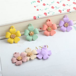 May include: A collection of colorful flower-shaped charms in yellow, green, peach, and purple. Each charm has a textured, gold-colored center and a loop for attaching to jewelry or crafts. The charms are arranged on a white surface.