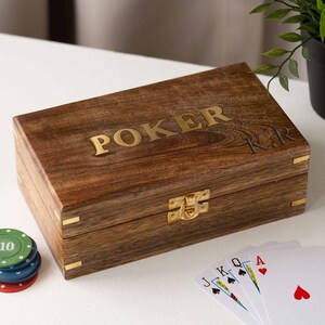 May include: A wooden poker box with the word "POKER" in gold lettering on the lid. The box has a gold clasp and corner accents. Poker chips and playing cards are visible next to the box.
