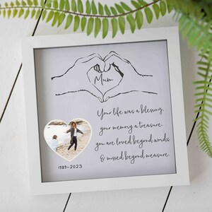 Puede incluir: Un marco de fotos blanco con fondo gris y un dibujo en blanco y negro de dos manos formando un corazón. La palabra "Mamá" está escrita dentro del corazón. El marco incluye una foto de una mujer y un niño caminando por la playa. El texto "Tu vida fue una bendición, tu memoria un tesoro. Eres amada más allá de las palabras y echada de menos más allá de toda medida. 1989-2023" está impreso debajo del dibujo.