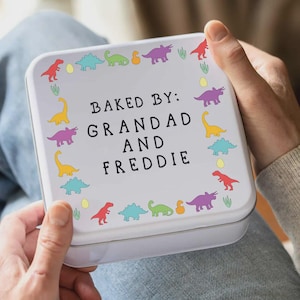 May include: A white, square tin with a lid, featuring colorful dinosaur illustrations around the border. The text "BAKED BY: GRANDAD AND FREDDIE" is centered on the lid. The tin is held by a person.