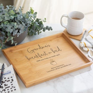 Pode incluir: Uma bandeja de caf&eacute; da manh&atilde; retangular de bambu com as palavras "Grandma's breakfast in bed" gravadas. A bandeja tamb&eacute;m tem o texto "Merry Christmas, Love from, all the grandkids xxx". Uma caneca, um porta-copos e um jornal tamb&eacute;m s&atilde;o vis&iacute;veis.