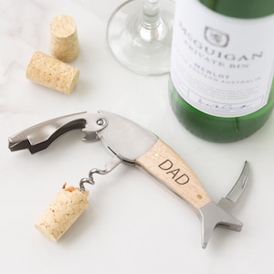 May include: A silver and wood corkscrew with a wooden handle that says "DAD" on it. The corkscrew is on a white surface with a wine bottle and corks.