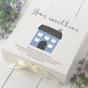 Pode incluir: Uma caixa de presente branca com uma ilustração de uma casa azul e cinza e o texto "Home sweet home" na tampa. A caixa é amarrada com uma fita branca e o texto "We are so proud of you. You have worked so hard and we can not wait for you to make this house and home. Love Mum & Dad" está impresso na caixa.