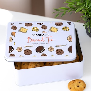 May include: A white rectangular biscuit tin with a lid decorated with cookie designs. The lid says "GRANDAD'S Biscuit Tin" in red script, with "NOT FOR ALFIE & SAMMY" underneath. The tin is open, showing a pile of chocolate chip biscuits.