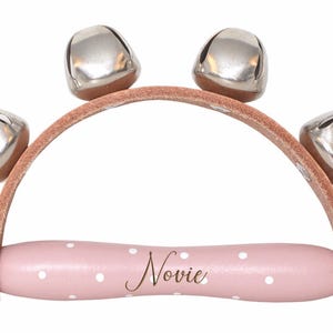 May include: A baby rattle with four silver bells attached to a light brown leather arch. The handle is pink with white polka dots and the name "Novie" in gold script.