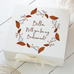 May include: White gift box with a brown floral wreath and the text "Bella. Will you be my Bridesmaid?"  The box is tied with a white satin ribbon.