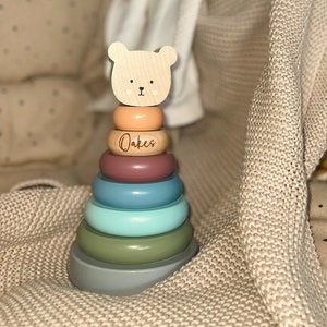 May include: A wooden stacking toy with a bear head and pastel-coloured rings. The rings are peach, mauve, blue, and green. The word "Oakes" is engraved on one ring. The base is grey. A classic toy for young children.