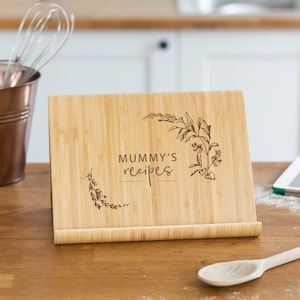 May include: A brown bamboo recipe stand with the text "MUMMY'S recipes" engraved on the front. The stand has a floral design and is angled to display a recipe book.