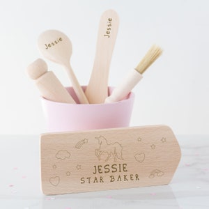 Puede incluir: Un letrero de madera con un unicornio, estrellas y un arcoíris. El letrero dice "Jessie Star Baker". También hay utensilios de cocina de madera en un tazón rosa.