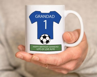 Op de afbeelding: Witte keramische mok met een ontwerp van een blauw en wit voetbaltenue. Het tenue heeft het woord "GRANDAD" en het nummer "1" erop. Onder het tenue bevindt zich een zwart-witte voetbal. De mok heeft een groene band aan de onderkant met de tekst "HAPPY BIRTHDAY GRANDAD LOTS OF LOVE ALFIE".