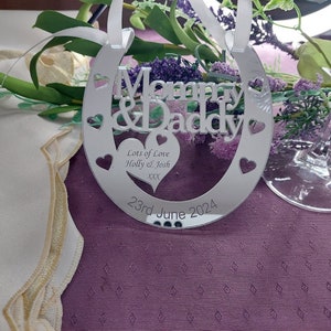 Pu&ograve; includere: Un ornamento a forma di ferro di cavallo argentato con le parole "Mommy & Daddy" e "Lots of Love Holly & Josh XX" e la data "23rd June 2024".