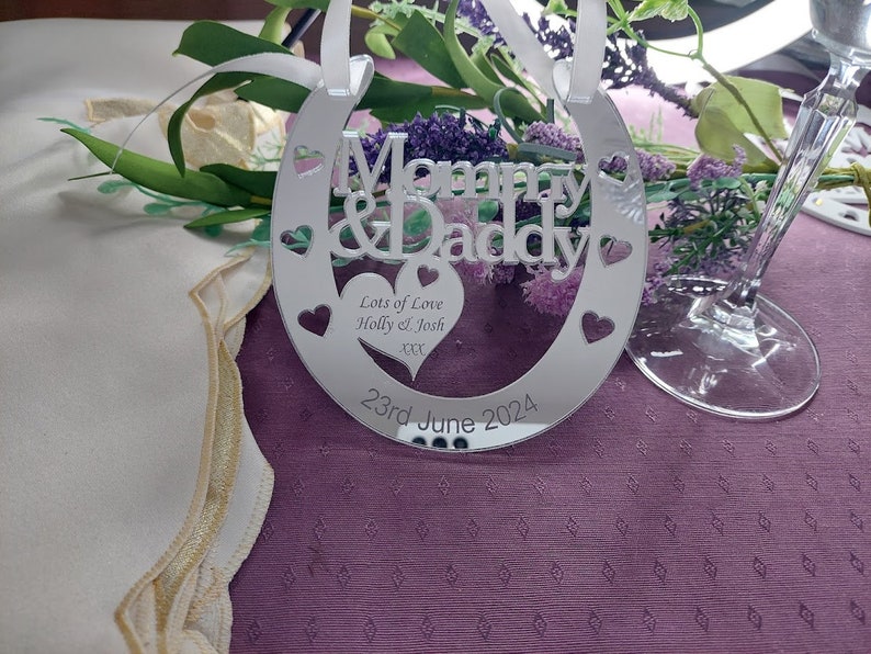 Pu&ograve; includere: Un ornamento a forma di ferro di cavallo argentato con le parole "Mommy & Daddy" e "Lots of Love Holly & Josh XX" e la data "23rd June 2024".