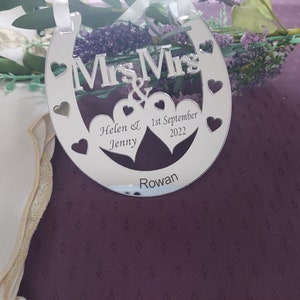 Puede incluir: Un adorno plateado en forma de herradura con las palabras "Mrs & Mrs" y dos corazones. El adorno est&aacute; personalizado con los nombres "Helen & Jenny", la fecha "1 de septiembre de 2022" y la palabra "Rowan".