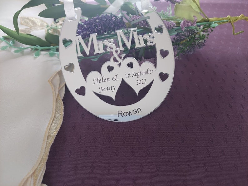 Puede incluir: Un adorno plateado en forma de herradura con las palabras "Mrs & Mrs" y dos corazones. El adorno est&aacute; personalizado con los nombres "Helen & Jenny", la fecha "1 de septiembre de 2022" y la palabra "Rowan".