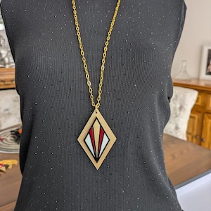 May include: A gold chain necklace with a diamond-shaped pendant. The pendant has a wooden frame with a white, red, and gold geometric design. The necklace is shown on a dark grey top.