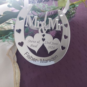 Puede incluir: Adorno plateado en forma de herradura con las palabras "Mr & Mr" y dos corazones. Los corazones tienen los nombres "Steve & Paul" y la fecha "23 de junio de 2024" inscritos en su interior. La parte inferior del adorno dice "Fisher-Marsden".
