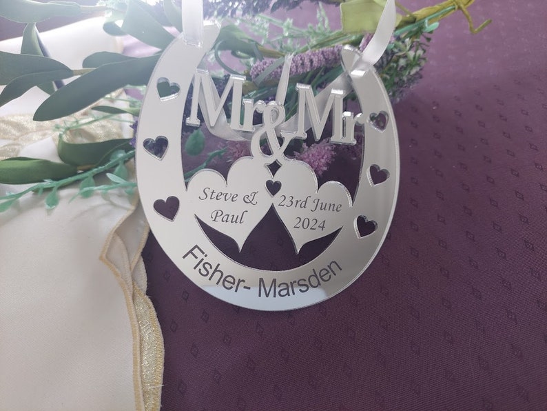 Puede incluir: Adorno plateado en forma de herradura con las palabras "Mr & Mr" y dos corazones. Los corazones tienen los nombres "Steve & Paul" y la fecha "23 de junio de 2024" inscritos en su interior. La parte inferior del adorno dice "Fisher-Marsden".