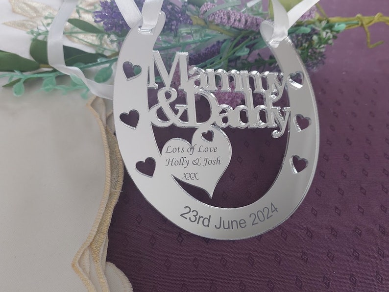 Pu&ograve; includere: Ciondolo a forma di ferro di cavallo argentato con le parole "Mommy & Daddy" e un cuore con il testo "Lots of Love Holly & Josh xxx" e la data "23rd June 2024".