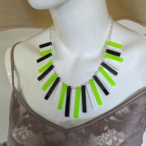 Puede incluir: Un collar hecho de piezas rectangulares negras, blancas y verde neón. El collar está en un maniquí blanco que lleva una blusa marrón.