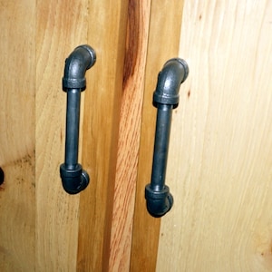 May include: Two black metal pipe-style door handles mounted on a wooden cabinet door.