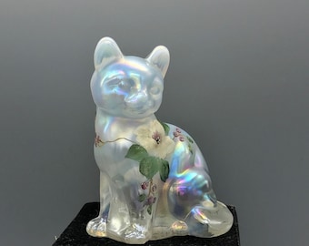 May include: Iridescent ceramic cat figurine with floral accents. The cat is seated, with a pearlescent finish that reflects multiple colors. The floral design includes white flowers, green leaves, and small purple berries.