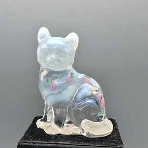 May include: A translucent glass cat figurine with a light blue body and white ears. The cat is decorated with pink and green floral accents. The figurine is sitting on a black base.