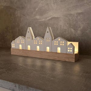 Op de afbeelding: Een wit keramisch dorpstaferel met huizen die van binnenuit verlicht zijn, op een houten basis. De huizen zijn in een rij geplaatst en hebben ramen en deuren.