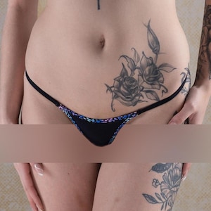 Puede incluir: Lingerie negra con un tatuaje floral en la cadera. La lencería tiene una tira fina y un delicado encaje.
