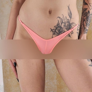 Puede incluir: Un tanga de color melocotón con tirantes finos. El tanga está hecho de una tela suave y elástica. El tanga lo lleva una persona con un tatuaje de rosa en la cadera.