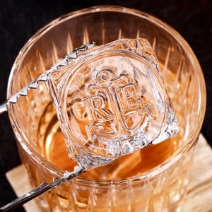 May include: A clear glass ice cube with an anchor and the letters "R.E." embossed on it. The ice cube is being held by a pair of tongs over a glass of amber liquid.