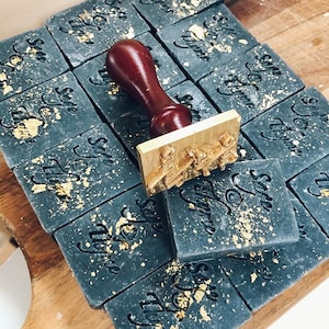 May include: A close-up of a wooden stamp with a brass base, used to imprint the words "Stay Wild" on black soap bars with gold flecks. The soap bars are arranged in rows on a wooden surface.