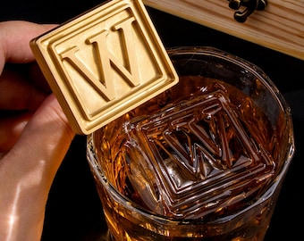 Op de afbeelding: Een vierkante goudkleurige metalen stempel met de letter W in een verhoogd schreeflettertype, vastgehouden naast een glas met amberkleurige vloeistof en een grote vierkante ijsblok met hetzelfde letter W-ontwerp.