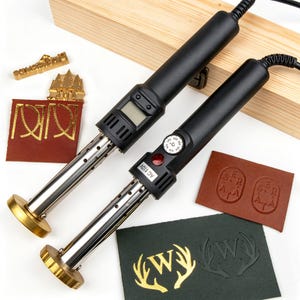 May include: Two black electric branding irons with metal heads, one featuring a digital display and the other a temperature dial labeled AC 110V. Included are several brass stamps and leather swatches showing embossed designs of letters and antlers.