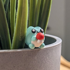 May include: A small, handmade polymer clay frog figurine in a light blue hue, holding a red mushroom. The frog has black eyes and a white belly. It sits in a gray concrete pot with green plant stems in the background.
