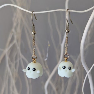 Op de afbeelding: Een paar zilveren en gouden oorbellen met witte spookbedels. De spoken hebben zwarte ogen en een kleine glimlach.
