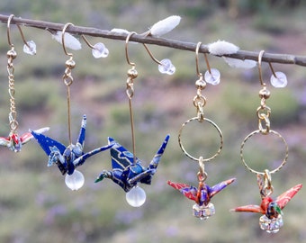 May include: A collection of origami crane earrings in various colors, including blue, red, and green, hanging from a branch. The earrings have gold-colored hooks and accents, with some featuring chain details and clear beads.