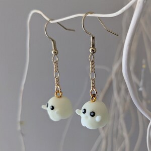 Op de afbeelding: Een paar oorbelletjes met een witte spookhanger met zwarte ogen. De spookhanger is bevestigd aan een gouden ketting en een zilveren haak.